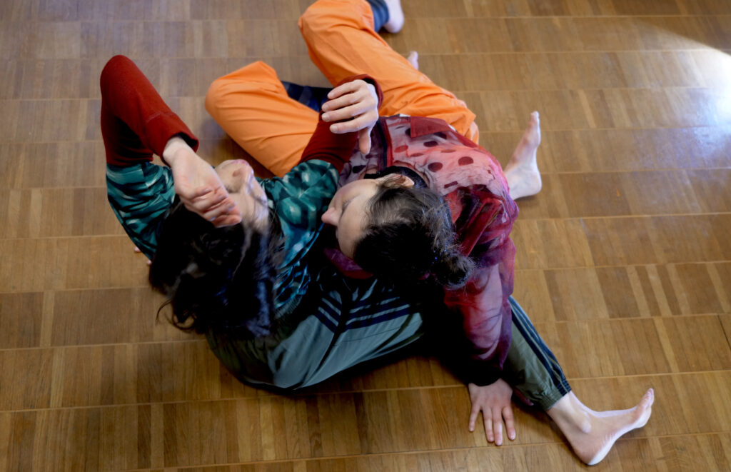 Zwei Menschen in Bunter Kleidrung bewegen sich auf einem Holzbodern. Sie bewegen sich übereinander mit ihren Körpern.